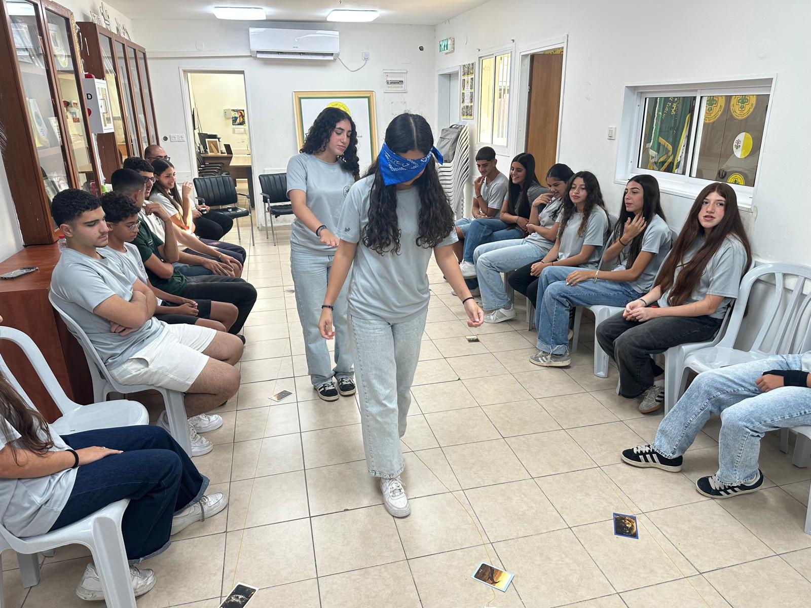A blindfolded participant walks between two rows of seated participants, guided by a peer, during a trust exercise.