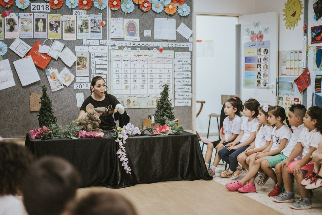 حلقةٌ صفّيّة: راويةٌ تستخدم الدُّمى وأطفالٌ منشدّون إليها، من زاويةٍ جانبيّة.