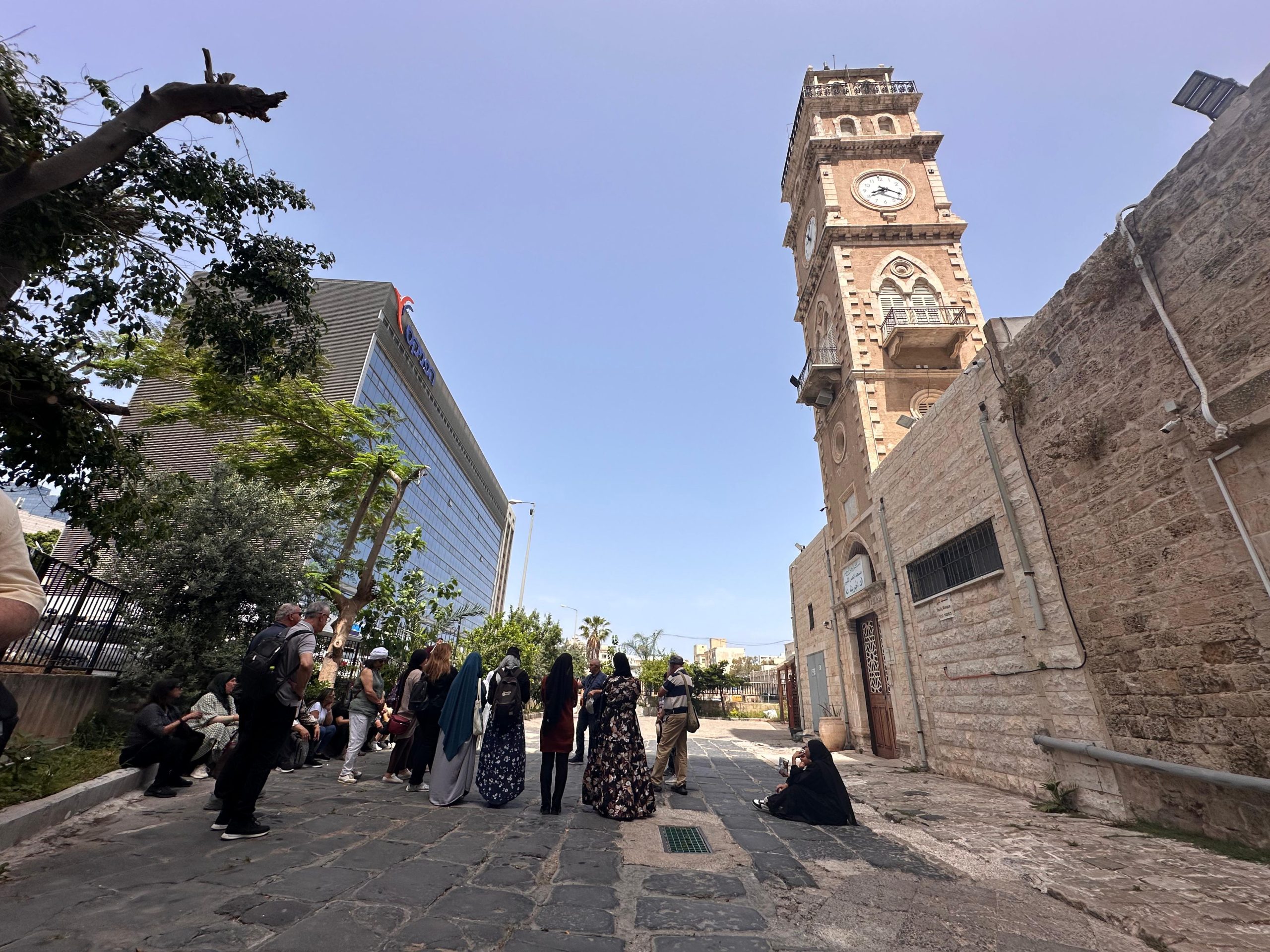 A community walk through the old city of Haifa, people seen mostly from behind, the clock tower above.