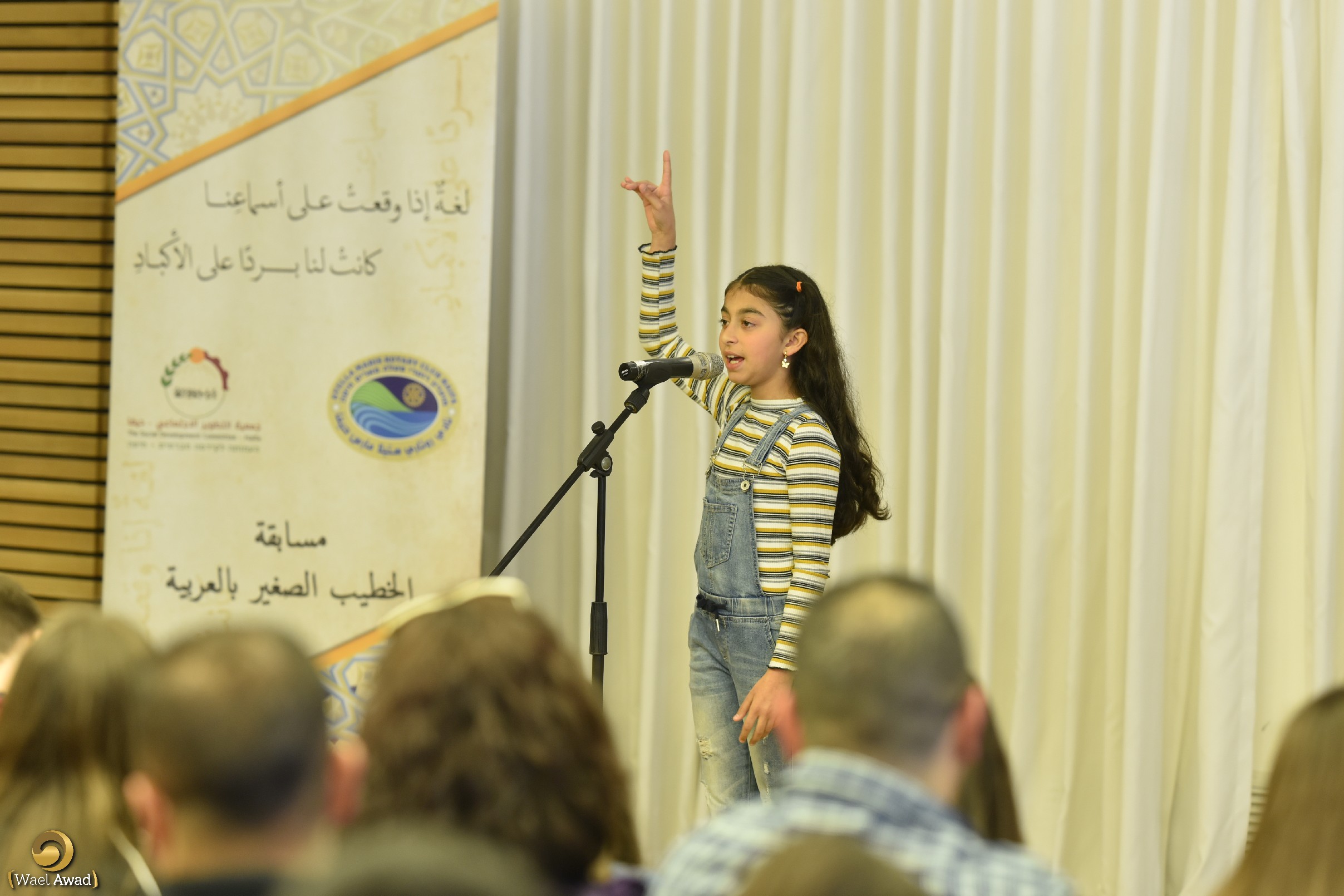 A girl at a microphone with her arm raised during an Arabic public-speaking contest.
